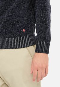 Pulover tricotat bleumarin, cu țesătură texturată, manșete și tiv ondulate. Prezintă o etichetă mică roșie pe partea stângă. Asortat cu pantaloni de culoare deschisă.