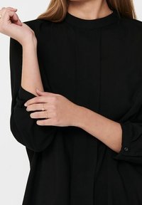 Tunique noire avec un col montant, des manches longues et des poignets à boutons, présentant une texture lisse et des plis sur le devant. Bague en or simple au doigt.