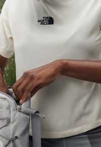 Kortärmad, krämfärgad skjorta med textilstruktur och svart logotyp. En person sträcker sig ner i en ljusgrå ryggsäck med dragkedjedetaljer.