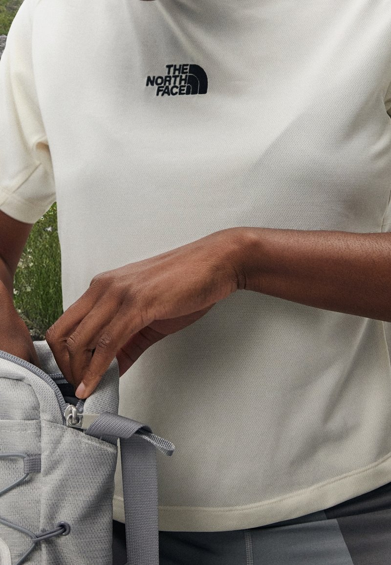 Kortärmad, krämfärgad skjorta med textilstruktur och svart logotyp. En person sträcker sig ner i en ljusgrå ryggsäck med dragkedjedetaljer.