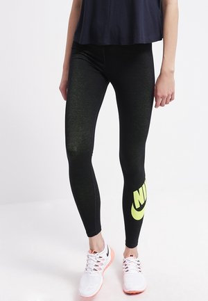 Osoba nosząca czarne legginsy Nike z dużym żółtym logo na lewej łydce oraz białe buty sportowe Nike z różowymi podeszwami.