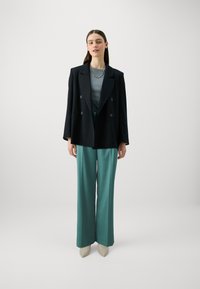 Svart dobbeltradet blazer over en teal topp og skreddersydde teal bukser. Spisse beige sko. Glitrende smykke detalj. Glatte stoffteksturer.