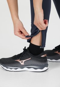 Svart idrottssko med roséguld-accenter, i kombination med marinblå tights med dragkedja i ärmslutet. Slät material och åtsittande design.