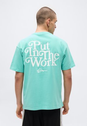 Homme aux cheveux courts de dos portant un t-shirt turquoise avec le texte blanc « Put In The Work » dans le dos sur un fond blanc uni.