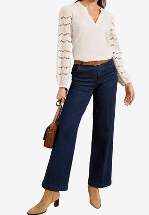 Femme portant un chemisier crème avec des manches en dentelle, un jean large taille haute foncé, une ceinture beige, des bottines beige et tenant un sac à main marron.