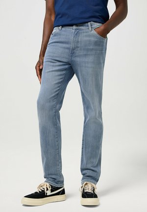 Jeans de mezclilla azul claro con pernera recta, diseño de cinco bolsillos y un leve desgastado. Llevados con zapatillas negras y una camisa azul marino.