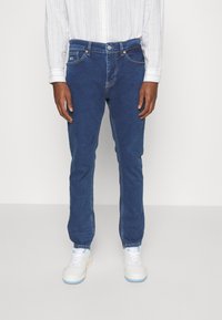 Blue denim jeans with a slim fit, five-pocket design, and subtle stitching. Paired with white sneakers and a light striped shirt.