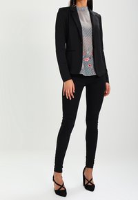 Svart skräddarsydd kavaj över en halvtransparent grå blommig topp. Slimmade svarta byxor och remmar svarta klackar fulländar outfiten.