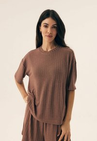 Brązowa ramiączkowa bluzka z krótkim rękawem o okrągłym dekolcie, charakteryzująca się luźnym krojem i detalem bocznej kieszeni. Gładka struktura materiału.
