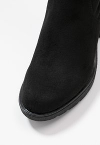 Bottine en suede noire avec un bout arrondi et une tige texturée. La semelle est plate et en caoutchouc, offrant adhérence et durabilité.