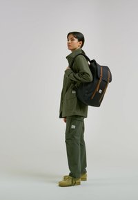 Black backpack with brown accents, worn by a person in an olive green jacket and pants. Soft texture, functional design, casual style.