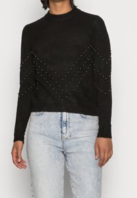 Czarny, dzianinowy sweter z okrągłym dekoltem, ozdobiony metalowymi ćwiekami ułożonymi w wzór zygzakowaty na rękawach i klatce piersiowej, połączony z jasnoniebieskimi dżinsami.