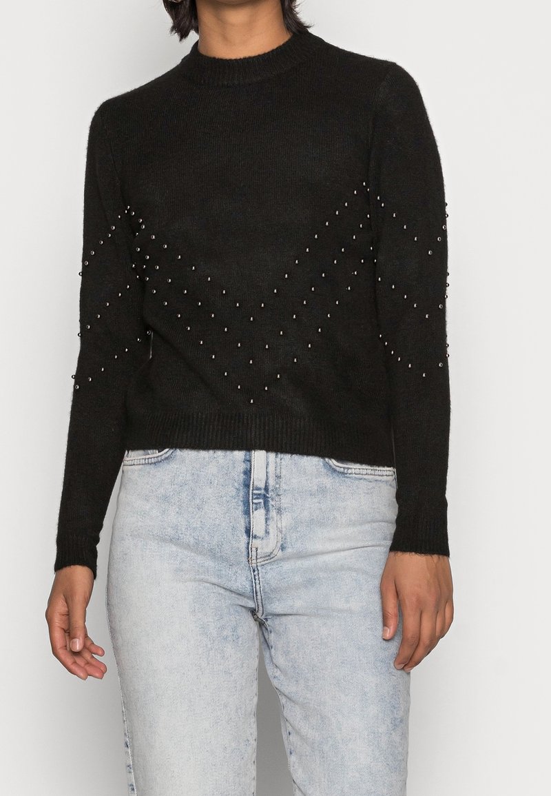 Czarny, dzianinowy sweter z okrągłym dekoltem, ozdobiony metalowymi ćwiekami ułożonymi w wzór zygzakowaty na rękawach i klatce piersiowej, połączony z jasnoniebieskimi dżinsami.