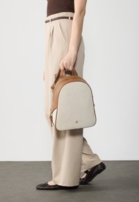 Mochila en tonos marrón claro y crema con una forma estructurada y redondeada. Presenta un frontal de tela texturizada y acentos de cuero suave. Se puede ver el tirador de la cremallera y el detalle del logo.