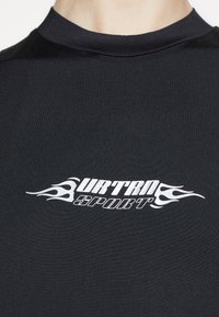 Schwarzes, eng anliegendes T-Shirt aus glattem Stoff mit einem weißen Flammenmotiv und dem Text "URTRA SPORT" in fetten Buchstaben über der Brust.