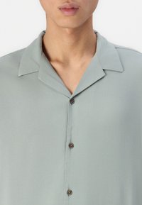 Cămașă scurtă cu manșete, de culoare verde deschis, fabricată dintr-un material neted, cu închidere prin butoane maro și o croială lejeră.