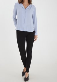 Blouse bleu clair avec un col, des manches longues et une coupe décontractée. Associée à un legging noir et des chaussures noires à bouts pointus. Texture lisse.