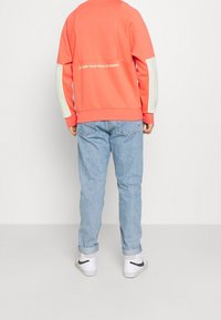Sweatshirt corail avec des manches vert clair, arborant un imprimé au dos "SURMONTEZ VOTRE PEUR DES HAUTEURS". Assorti avec un jean clair à revers et des baskets blanches.