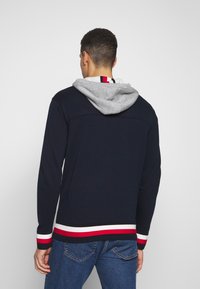 Veste à capuche zippée marine avec une capuche grise, des poignets côtelés et un ourlet rayé en blanc, rouge et marine. Portée par-dessus une chemise blanche avec des accents colorés.