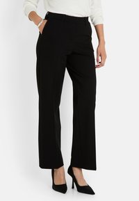 Mujer vestida con pantalones rectos negros y tacones altos negros, con una mano en el bolsillo y una blusa blanca de manga larga visible.