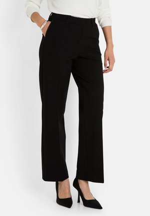 Femme portant un pantalon noir à jambes droites et des talons hauts noirs, avec une main dans la poche et un haut à manches longues blanches visible.