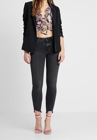 Blazer nero sopra un crop top con motivo rosa e nero. Jeans neri aderenti con orli sfrangiati, abbinati a tacchi a strisce rosa e bianchi.