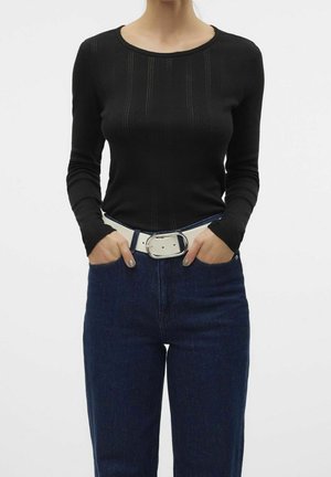 Vrouw die een nauwsluitende zwarte top met lange mouwen draagt, ingestopt in een hooggesloten donkere blauwe spijkerbroek met een brede witte riem, handen in de zakken.