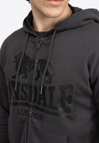 Šedá mikina na zips so grafikou leva, s kapucňou na šnúrku a prednými vreckami. Vyrobená z mäkkej tkaniny s hladkou textúrou.