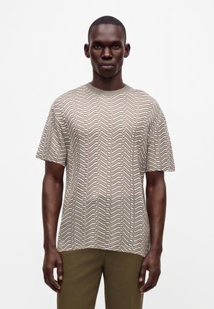 Uomo che indossa una maglia a maniche corte beige e marrone con motivo a zigzag e pantaloni verde oliva, in piedi su uno sfondo bianco semplice.