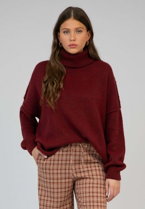 Jeune femme aux longs cheveux bruns portant un pull à col roulé bordeaux et un pantalon beige à carreaux, debout devant un fond uni.