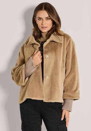 Vrouw met een lichtbruine nepbonten jas over een beige gebreide trui en zwarte broek, staand tegen een effen lichte achtergrond.