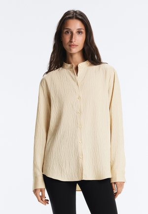 Donna con capelli castani lunghi che indossa una camicia beige strutturata con bottoni e collo alla coreana e pantaloni neri, in piedi davanti a uno sfondo bianco.
