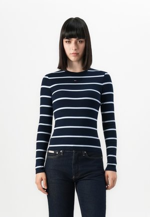 Jeune femme avec des cheveux sombres et raides, vêtue d'un t-shirt à manches longues bleu marine avec des rayures blanches horizontales et d'un jean foncé, se tenant contre un fond blanc.