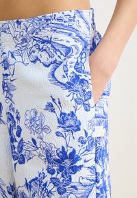 Blommönstrade byxor i blått och vitt, med en texturerad tyg, elastisk midja och sidoficka. Mönstren inkluderar olika blommor och blad.