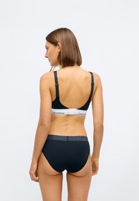Černá bezdrátová podprsenka se širokým bílým pásem s logem "Calvin Klein", doplněná černými kalhotkami se šedým páskem. Hladký povrch.