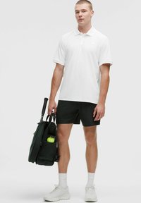 Biela polo tričko, čierne šortky a biele tenisky. Osoba má tmavozelenú tašku s viditeľným tenisovým loptičkom vo vnútri.