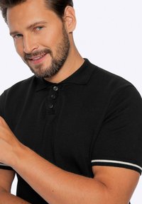 Černé polo tričko s texturovaným povrchem, tříknoflíkovou légou a bílými akcenty na rukávech. Model se usmívá.