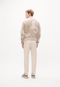 Man die met zijn rug naar de camera staat, gekleed in een beige gebreide trui, lichtcrème broek en beige sneakers, tegen een plain witte achtergrond.