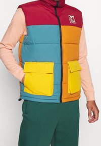 Barevná péřová vesta s červenými, modrými, oranžovými a žlutými částmi. Má vysoký límec a dvě žluté kapsy s klopami. Texturovaná látka.