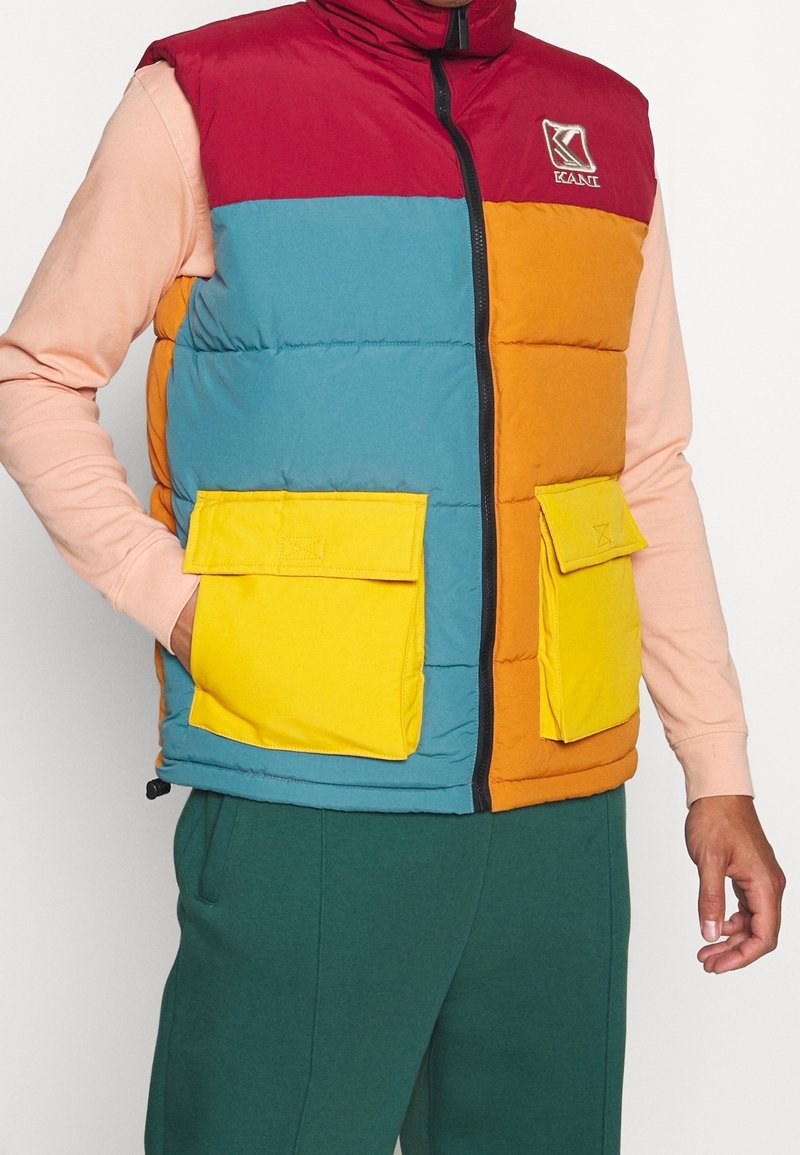 Barevná péřová vesta s červenými, modrými, oranžovými a žlutými částmi. Má vysoký límec a dvě žluté kapsy s klopami. Texturovaná látka.