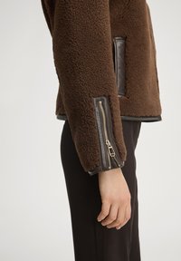 Brązowa kurtka polarowa z ozdobami ze skóry, z suwakiem na mankiecie. Tekstura jest miękka i puszysta, o pudełkowatym kroju.