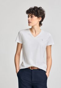 Vit v-ringad t-shirt i mjuk bomull, med korta ärmar och diskret logotypbroderi på vänster bröst. Bärs med marinblå byxor och ett brunt bälte.