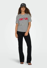 Gestreept zwart-wit T-shirt met een vet rood "AMOUR"-opdruk, gecombineerd met zwarte flared jeans en een grijze pet.