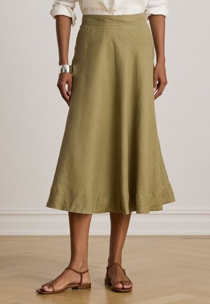Femme portant une jupe midi droite vert olive, une chemise blanche aux manches longues retroussées, un bracelet manchette argenté et des sandales plates marron.