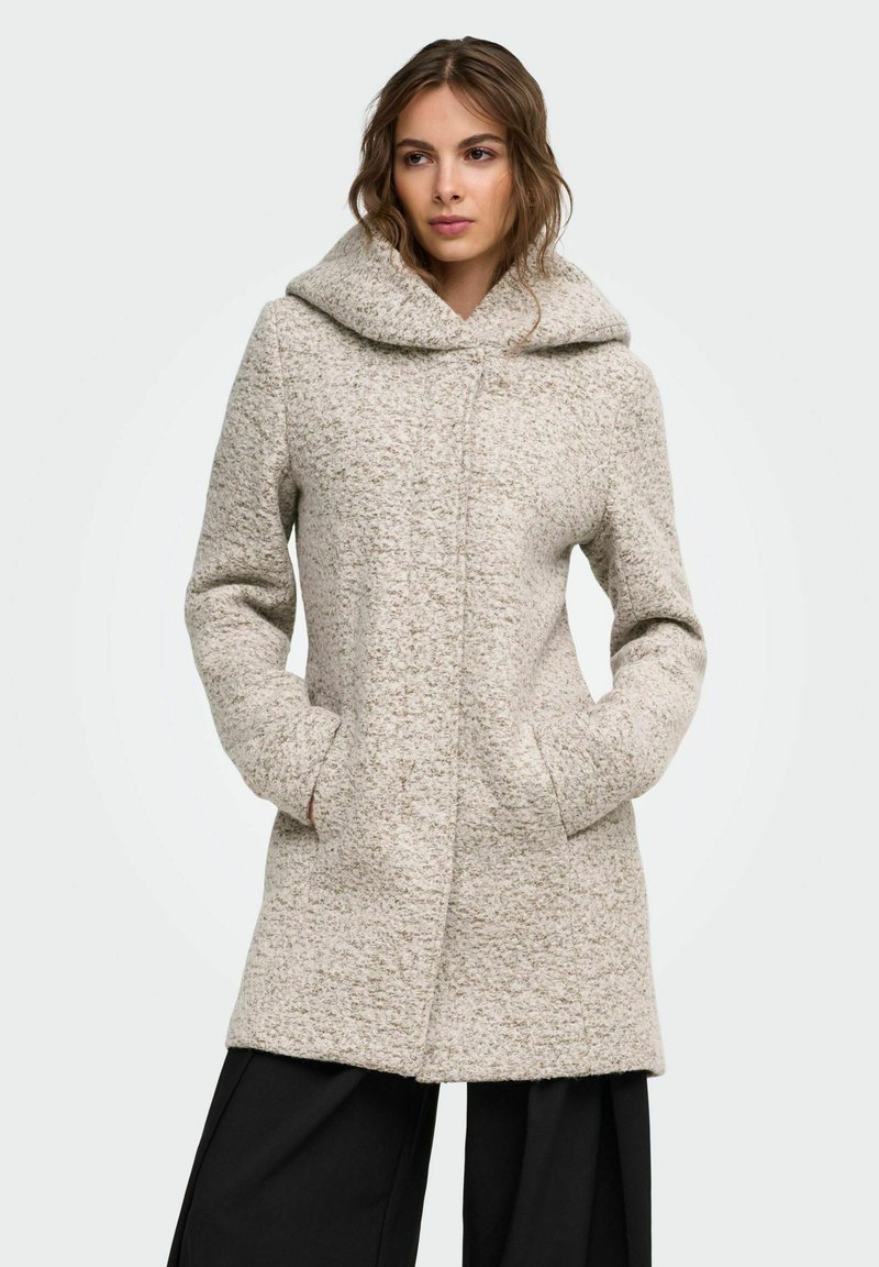 Femme portant un manteau texturé gris clair avec un large col et les mains dans les poches, associé à un pantalon noir ample, regardant légèrement sur le côté.