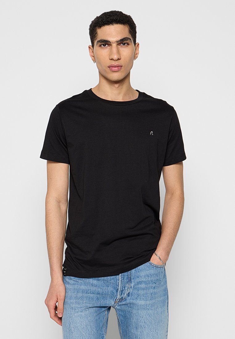 T-shirt en coton noir avec un col rond et des manches courtes. Présente un petit logo sur la poitrine et une coupe décontractée. Porté avec un jean bleu.