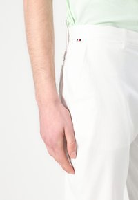 Białe spodnie wykonane z gładkiego materiału, z boczną kieszenią, płaskim przodem oraz małą metką z logotypem w czerwono-białoniebieskich kolorach na pasku.