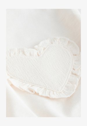 Vaaleanbeige kangas, jossa sydämen muotoinen, teksturoidulla pinnalla ja röyhelöisillä reunoilla varustettu paikka, valkoista taustaa vasten.