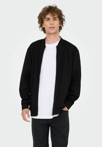 Sort zip-up bomberjakke med ribbede cuff og krave, iført over en hvid T-shirt, parret med mørke jeans. Ensartet baggrund.