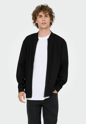Chaqueta bomber negra con cremallera, puños y cuello de canalé, usada sobre una camiseta blanca, combinada con pantalones vaqueros oscuros. Fondo liso.